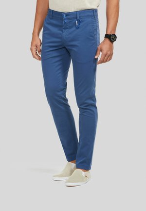 Man met slim fit blauwe broek met knoop en rits, blauwe karabijnhaak aan de lussch om de riem, zwart horloge en beige instapschoenen.