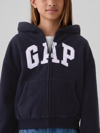 Jovem a usar um hoodie com fecho azul-escuro, em fleece, com grandes letras brancas "GAP" no peito e bolsos frontais.