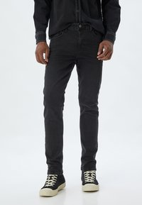 Jeans de mezclilla negra con corte ajustado, que cuentan con cierre de botón y cinco bolsillos. Llevados con zapatillas negras de caña alta y suelas blancas.