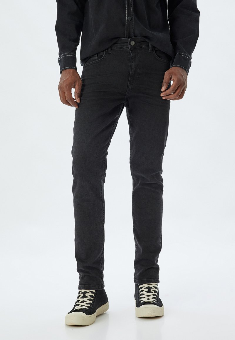 Jeans de mezclilla negra con corte ajustado, que cuentan con cierre de botón y cinco bolsillos. Llevados con zapatillas negras de caña alta y suelas blancas.