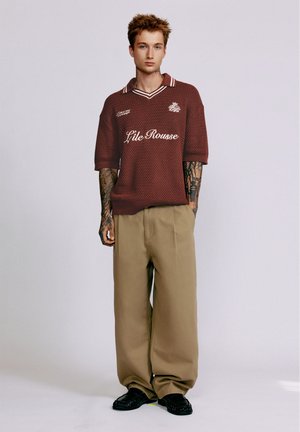 Jeune homme aux cheveux courts et tatouages, portant un pull en maille bordeaux avec un texte et un pantalon large beige sur un fond uni.