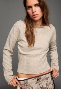 Világos bézs színű kötött pulóver hosszú ujjakkal és bordázott alsó résszel. Az anyag puhanak tűnik, enyhe textúrával, amit mintás nadrág egészít ki.