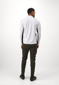 Witte langemouwen shirt met zwarte zijpanelen, gecombineerd met olijfgroene pantalons en zwarte sportschoenen. Soepele stof, getailleerde pasvorm.