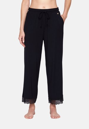 Černé široké kalhoty s elastickým páskem na stažení a krajkovou lemovkou na nohavicích, model stojí bosý s jednou rukou v kapse.