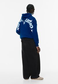 Kongeligt blå hoodie med hvid skrift og stjernesymboler på ryggen, kombineret med løse sorte denimbukser og beige fodtøj.