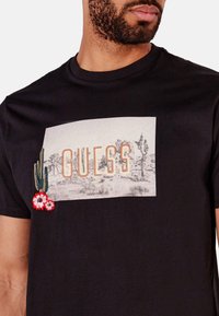 Camiseta negra con un diseño gráfico que incluye la palabra "GUESS" y acentos bordados de cactus y flores en colores vibrantes.