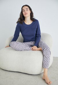 Mörkblå långärmad topp med en ficka, kombinerad med mönstrade ljusa byxor med ett zigzag-mönster. Modell sitter på en texturerad, rund stol.
