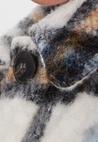 Wollstoff in Weiß, Grau und Braun mit einem karierten Muster. Verfügt über einen schwarzen Knopf und eine sichtbare, weiche Textur mit flauschigen Fasern.