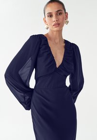 Námořnicky modré šaty s hlubokým výstřihem do V, volánkovým živůtkem a dlouhými průsvitnými rukávy. Hladká textura a přiléhavý střih. Minimalistický design.