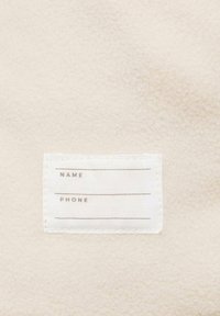 Krämfärgad fleece med en mjuk textur. En vit tygskylt har blanka linjer för "NAMN" och "TELEFON" i subtil grå tryck.