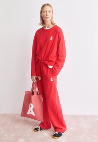 Rode katoenen lange mouwen shirt en bijpassende joggingbroek met wit lintlogo, gecombineerd met een roze draagtas met een soortgelijk logo en zwarte sneakers.