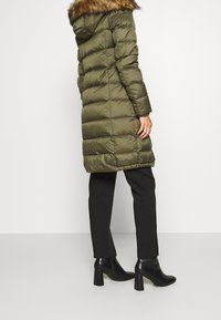 Cappotto verde oliva imbottito con cappuccio e finiture in pelliccia sintetica, caratterizzato da trapuntatura orizzontale e lunghezza fino al ginocchio. Indossato con pantaloni neri e stivali con tacco.