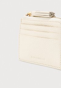 Kreemi nahast krediitka hoidja, millel on mitu sloti, kuldne lukuga ja dekoratiivne pom-pom tõmbepeaga, valge tausta peal.