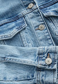 Tejido de denim azul claro con textura visible, que presenta un cierre de botón y acentos cosidos. Dos botones están grabados con "NUEVAS PANTALONES DESDE 1968."
