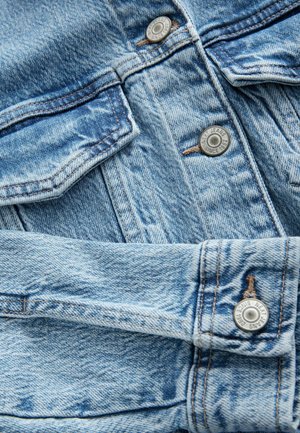 Heledsinise denimkangas, millel on nähtav tekstuur, nuppude sulgemise ja õmmeldud detailidega. Kaks nuppu on pressitud kirjadega "Uued teksad alates 1968."