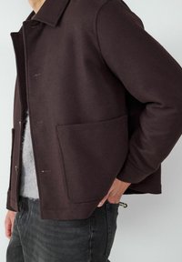 Ciemnobrązowa kurtka z miękkiego materiału, z kołnierzem, guzikiem z przodu i dużymi bocznymi kieszeniami. Nosi się ją na szarym ubraniu.