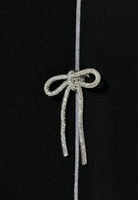 Prenda negra con un broche de lazo de strass, con dos tiras colgantes y un ribete plateado al lado del cierre. Textura suave.