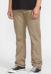 Chinos de algodón beige con un corte relajado, que presentan bolsillos laterales, una textura suave y se combinan con zapatillas de lona negras.
