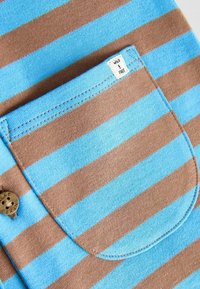 Gros plan d’un tissu rayé bleu et marron avec une poche cousue et une petite étiquette indiquant « WILD & FREE ».