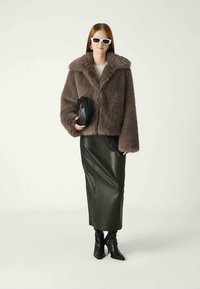 Donna che indossa occhiali da sole bianchi, giacca in pelliccia artificiale marrone, gonna midi in pelle nera, stivali con tacco neri, mentre tiene una borsa a clutch nera.