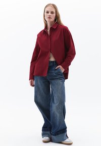 Rotes Button-Up-Hemd mit lockerer Passform und abgerundetem Saum, kombiniert mit weit geschnittenen blauen Jeans und weißen Sneakers.