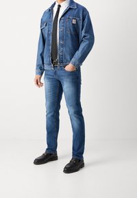 Giacca di jeans blu con taschino sul petto, abbinata a jeans azzurri chiari. Mocassini neri con un accento dorato. Sotto, una semplice t-shirt grigia.