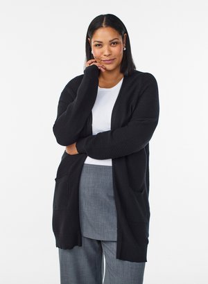 Ung kvinde med glat sort hår iført hvid top, sort cardigan og grå bukser, stående med hånden på hagen foran en hvid baggrund.