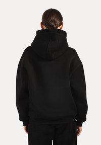 Personne aux cheveux foncés attachés, portant un sweat à capuche noir surdimensionné et un pantalon noir, debout de dos sur un fond uni.