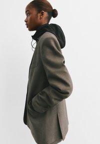 Blazer marron texturé avec une couche à capuche noire en dessous. Le blazer a une poche unique et des boutons aux manches. Vue de profil.