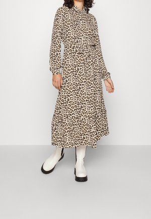 Femme portant une robe midi à manches longues à imprimé léopard beige et marron avec ceinture nouée à la taille, associée à des bottes blanches épaisses sur un fond uni.