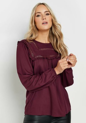Blouse longue bordeaux avec un col à volants et des coutures décoratives. Tissu lisse, coupe décontractée et poignets froncés.
