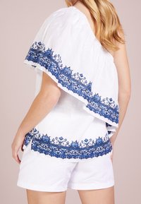 Vit enaxlad topp med blå blommig broderi längs fållen och ärmen, tillverkad av lättviktig tyg, kombinerad med vita shorts.