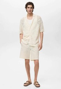 Brede crèmekleurige cardigan van breiwerk met open structuur en korte mouwen, gecombineerd met lichtcrèmekleurige shorts en bruine sandalen, wat een casual zomerse look creëert.