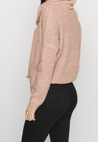 Rosafarbener, grob gestrickter Pullover mit hohem Kragen, lockerem Schnitt und langen Ärmeln, kombiniert mit eng anliegenden schwarzen Hosen. Detail der strukturierten Stoffstruktur sichtbar.