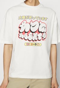 Bílá tričko s nadměrným japonským textem v žluté, bílé a černé, obtažené červenou, nošené osobou v béžových kalhotách.