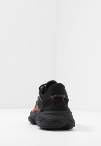Černá sportovní obuv s červenými akcenty, vyrobená z síťoviny a gumy. Má texturovanou podrážku a kulatý kovový detail na patě.