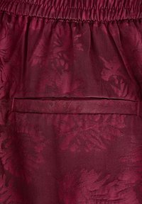 Burgunderfärgad tyg med en texturerad blommönster, utrustad med elastisk midja och en horisontell fickdetalj.