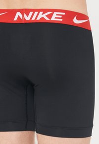 Șorturi sportive negre cu un brâu elastic roșu, având logo-ul Nike în alb. Textură netedă și design ajustat.