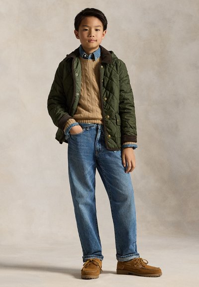 Jongen draagt een groene doorgestikte jas, een beige kabelgebreide trui, een blauw denim overhemd, spijkerbroek en bruine veterschoenen, staand met één hand in de zak.