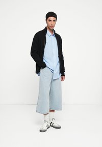 Mann i svart cardigan, blåstripete skjorte, vide lyse denimbukser, hvite sokker og hvite joggesko som står mot en hvit bakgrunn.