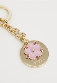 Llave metálica dorada con un colgante floral. Detalle de flor de esmalte rosa en un charm redondo que presenta el texto grabado "Furla". Diseño de cadena texturizada.