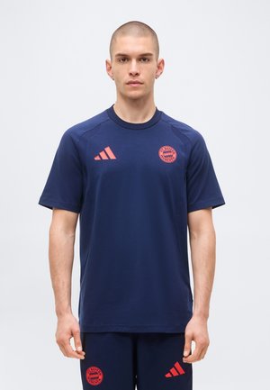 Mann steht in dunkelblauer Sportbekleidung des FC Bayern München mit roten Logos auf Shirt und Shorts vor einfachem hellem Hintergrund.