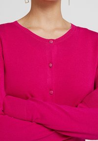 Fuksianvärinen neulotty neuletakki pyöreällä pääntiellä, viisi nappia edessä ja resorireunaiset hihansuut. Pehmeä pinta ja tasaiset saumat.