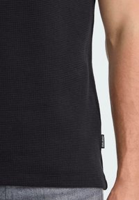 Fekete, textúrált ujjatlan felső, méhsejt mintával, hem címkével. Az anyag könnyűnek és légáteresztőnek tűnik. Látható váll és kar.