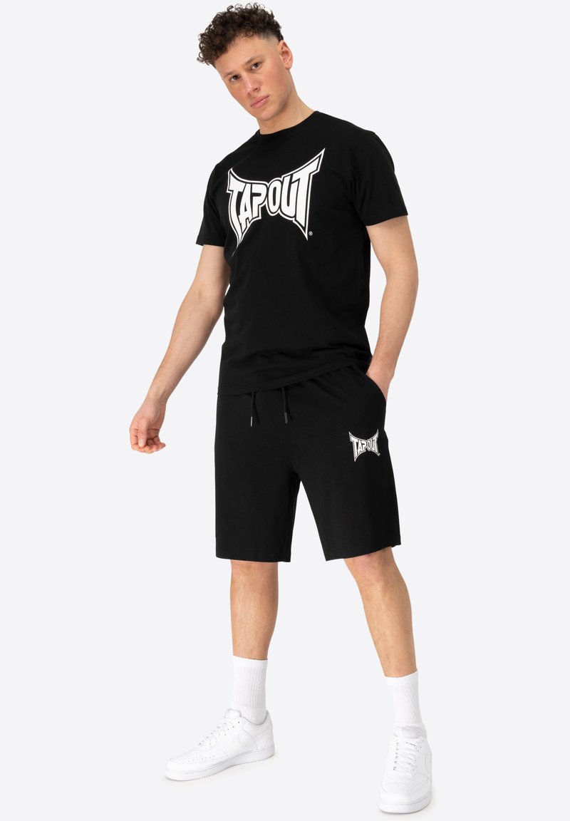 Melnā kokvilnas T-kreklā ar lielu baltu "TAPOUT" logotipu, apvienots ar melnām šortiem ar to pašu logotipu. Valkāts ar baltām sporta apaviem un zeķēm.