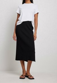 Hvid t-shirt med korte ærmer parret med en sort ribbet midi-nederdel, der har en sidelomme. Sorte flade sandaler fuldender outfittet.
