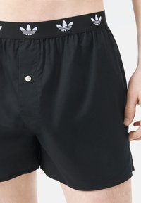 Svart herrshorts med knappstängning och elastisk midja med vita Adidas trefoil-logotyper. Lätt tyg med en mjuk textur.