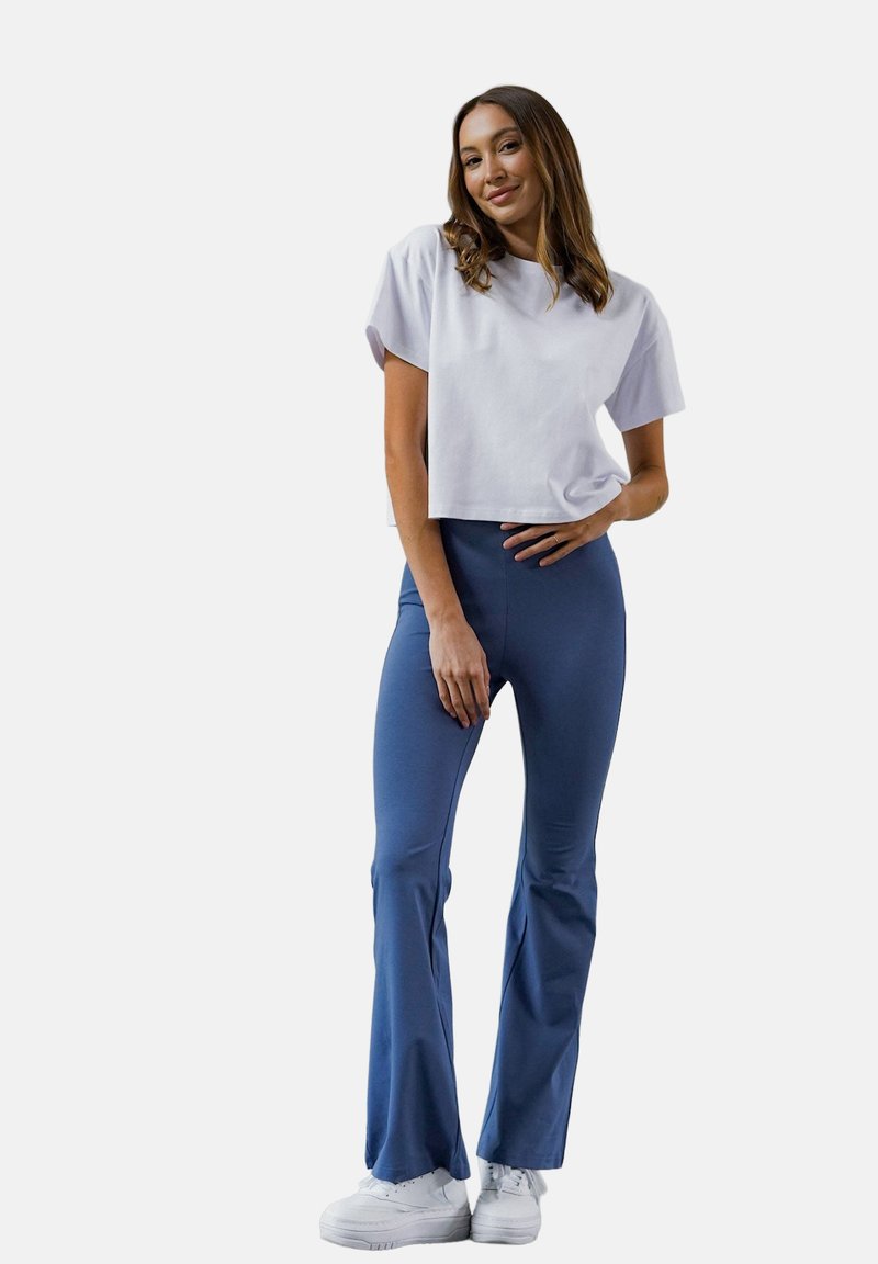 Przycięta biała koszulka z krótkimi rękawami, w połączeniu z niebieskimi legginsami dzwonami. Modelka nosi białe trampki, a tkanina jest gładka w dotyku.