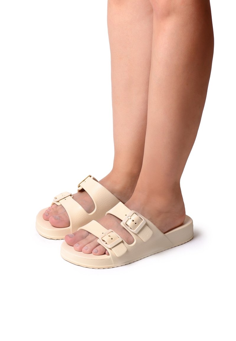 Pieds portant des sandales beige à enfiler avec deux brides à boucles réglables, présentés sur un fond blanc.