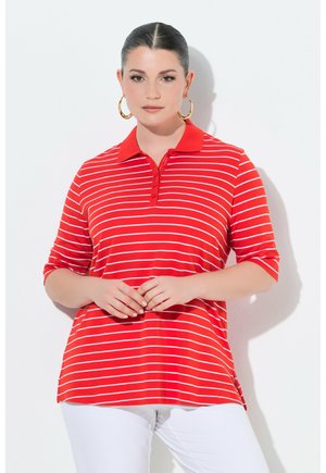 Donna con capelli lisci tirati indietro che indossa orecchini a cerchio dorati, una maglietta polo a righe rosse e bianche e pantaloni bianchi, in piedi contro uno sfondo bianco.